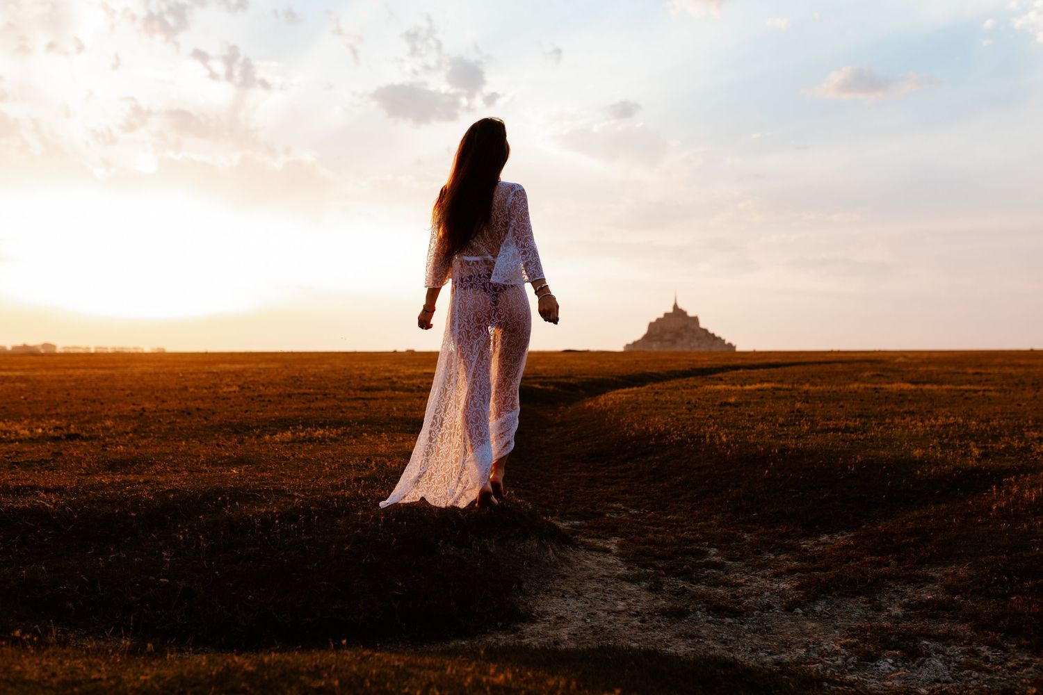 Woman in white dress walks toward Mont Saint-Michel at golden sunset across open fields.