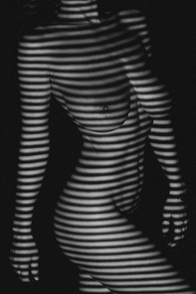 Black and white artistic nude photograph with striped shadow patterns creating abstract lines across a female silhouette.