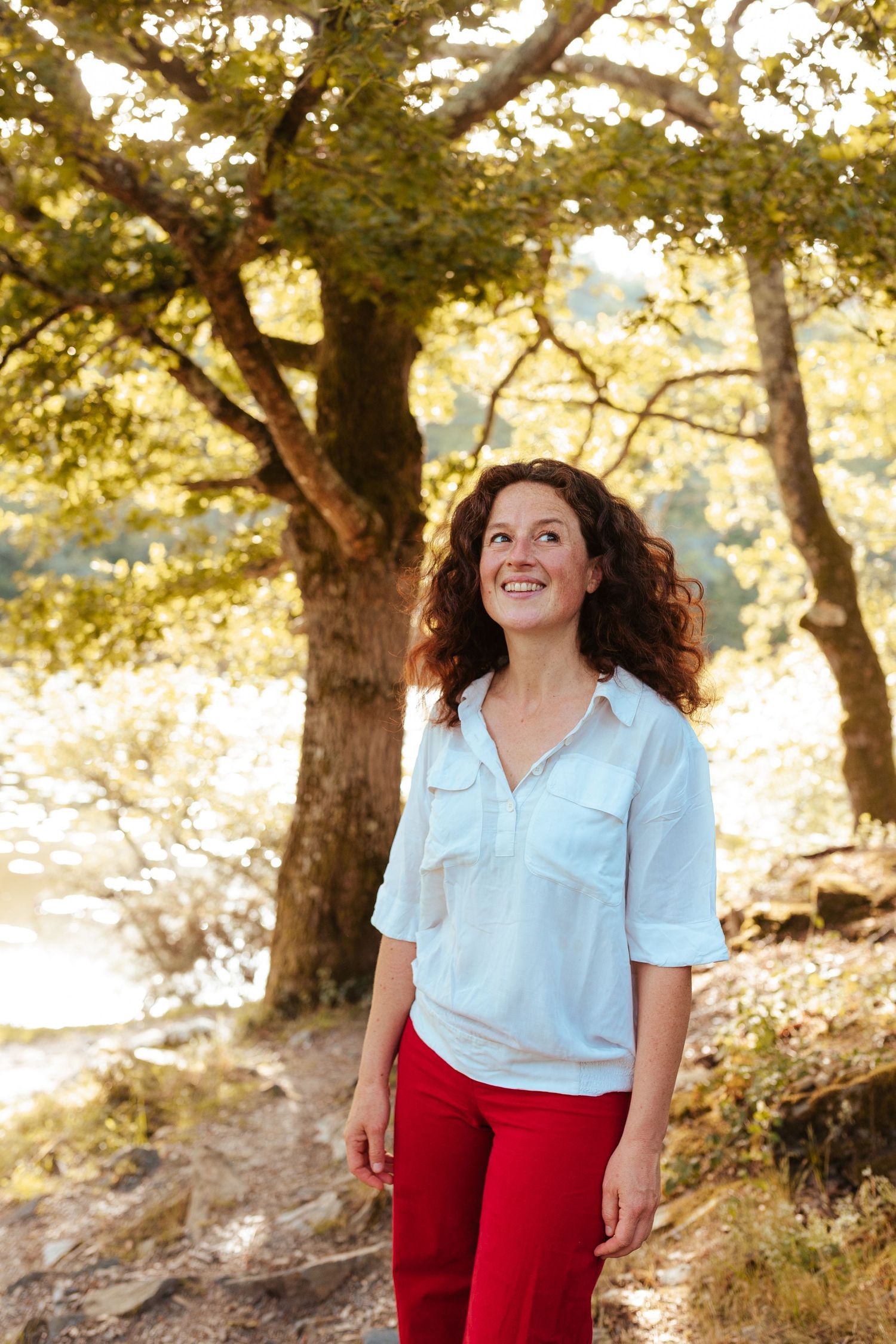 A smiling woman with curly hair in a white blouse and red pants stands outdoors near trees by a river.