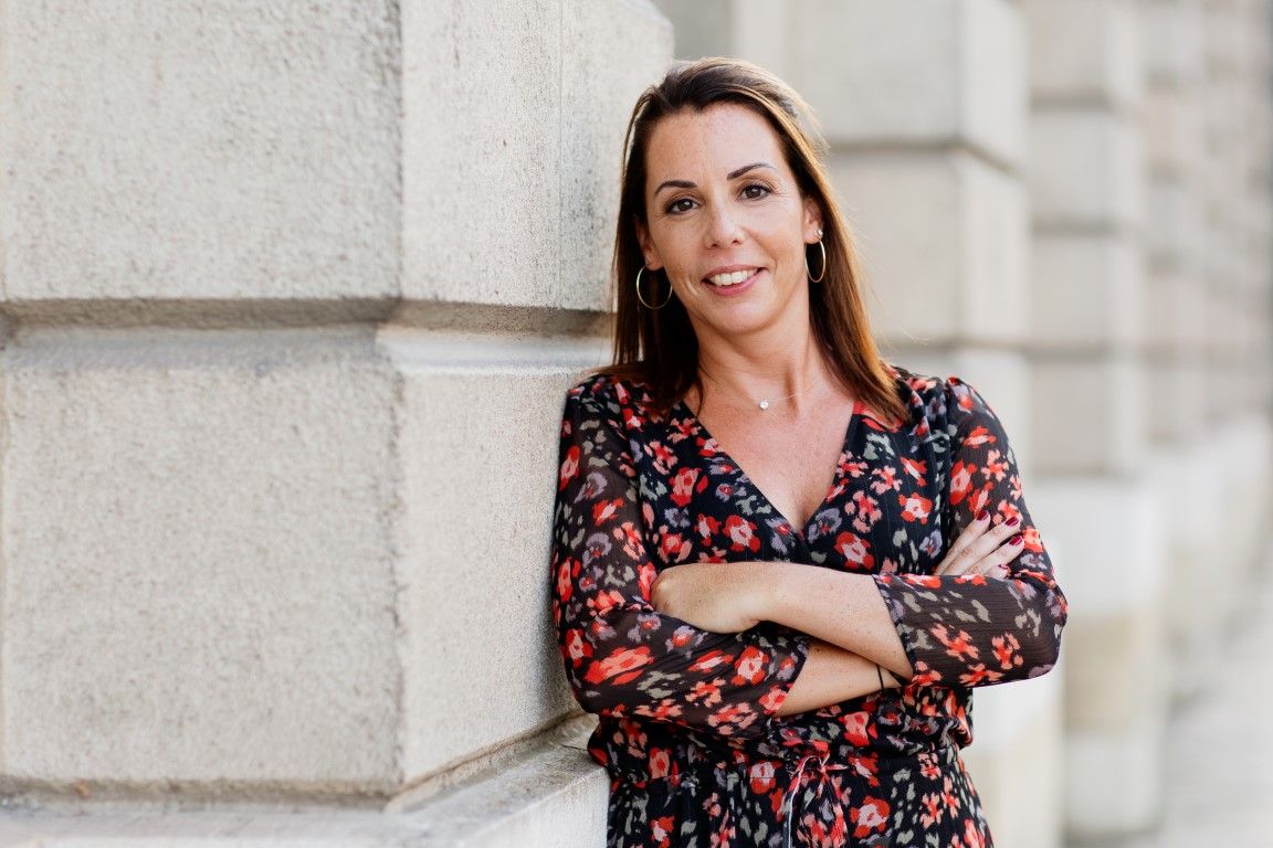Smiling woman in floral dress leaning against stone column with arms crossed outdoors.