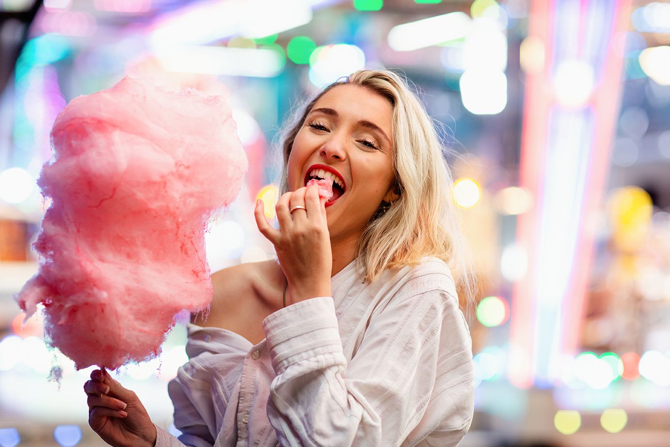 Une femme blonde souriante mange de la barbe à papa rose dans une fête foraine colorée, de nuit.