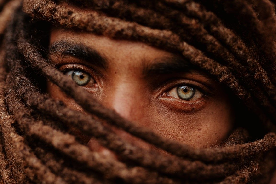 Close-up of piercing green eyes framed by brown dreadlocks wrapped around a face.