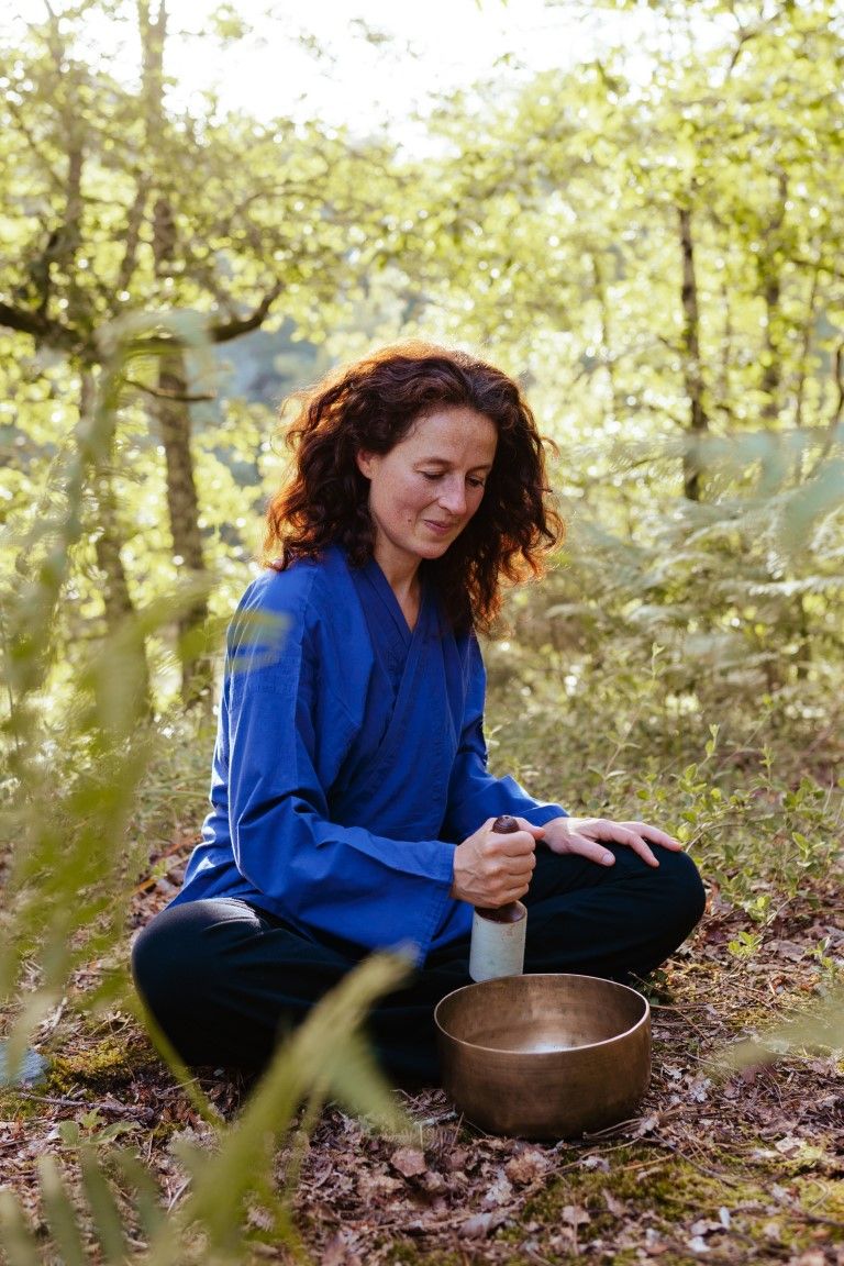 Woman in blue top meditating outdoors in forest with singing bowl, practicing mindfulness in nature.