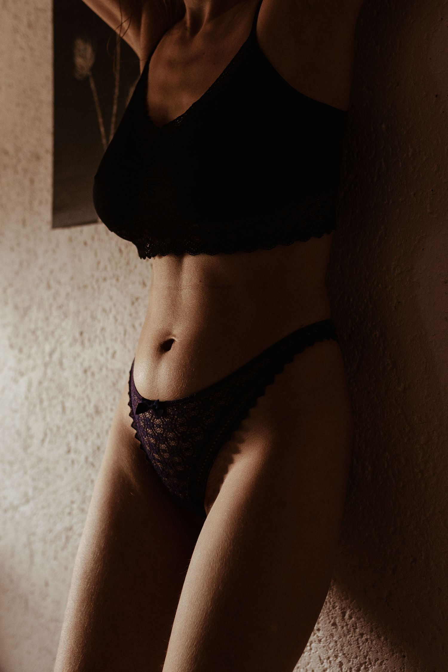 Femme en soutien-gorge en dentelle noire et string posant devant un mur texturé sous un éclairage dramatique.