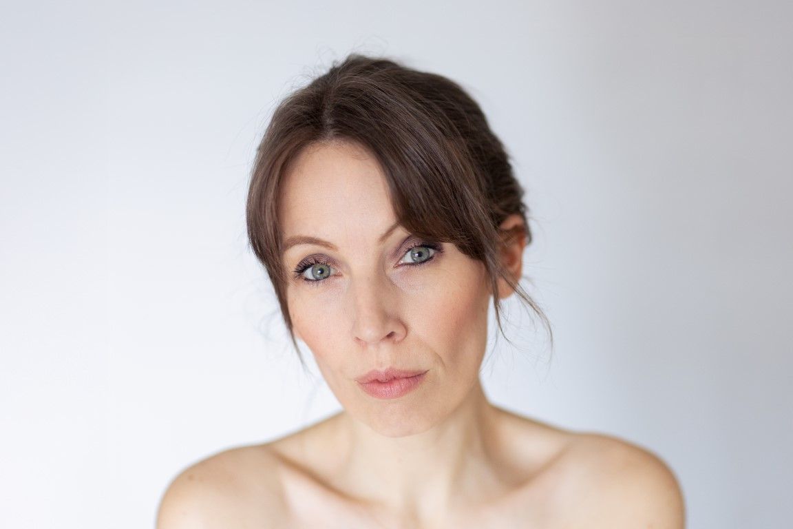 Portrait en gros plan d'une femme brune aux yeux bleus et au maquillage naturel sur fond gris clair.