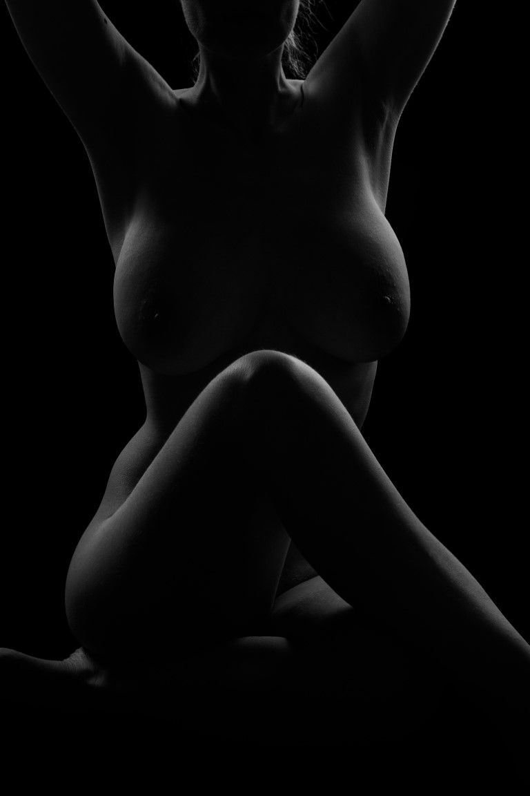 Black and white artistic nude silhouette of a pregnant woman kneeling against a dark background.