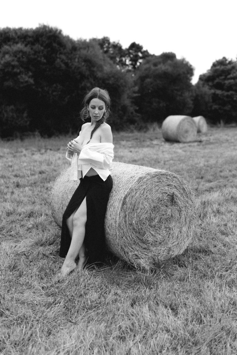 Black and white photo of a woman in a white shirt and black skirt leaning against a hay bale in a field.