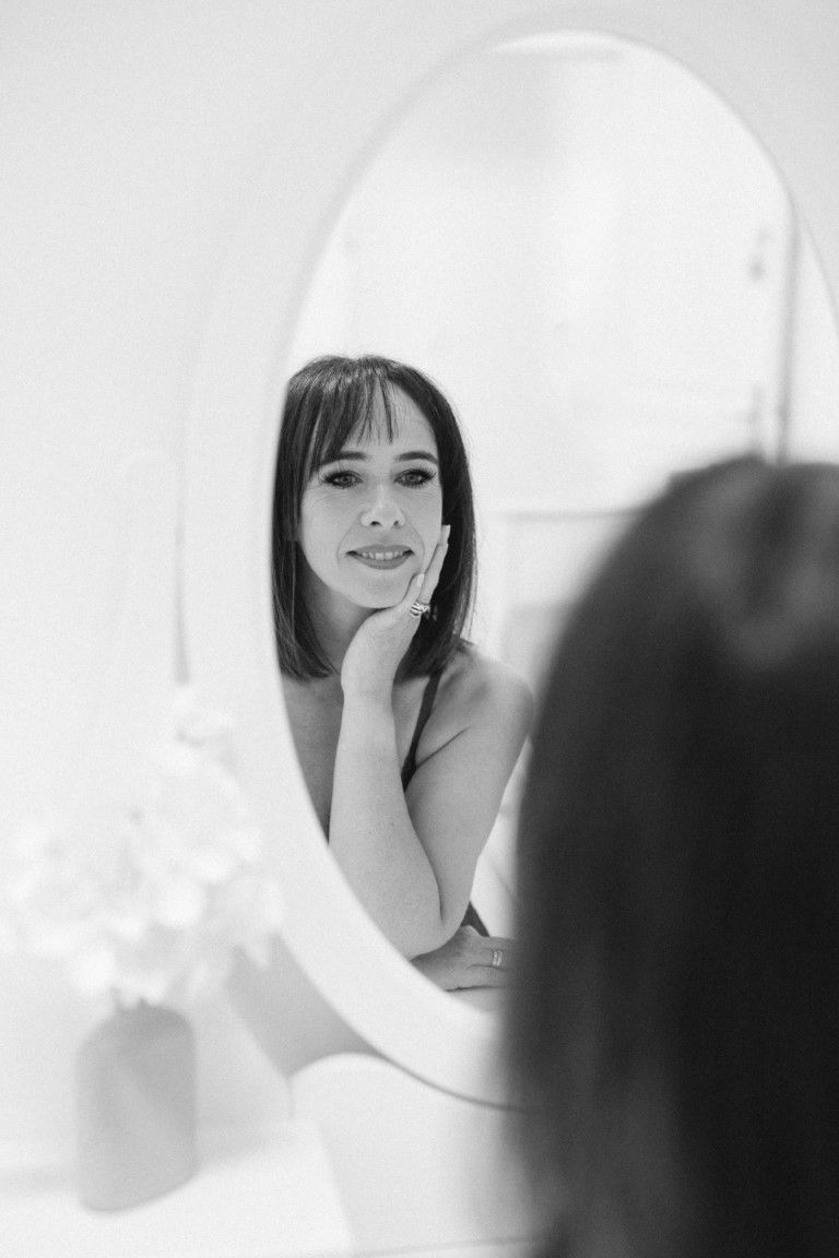 Black and white portrait of a brunette woman with bangs posing by a round mirror.
