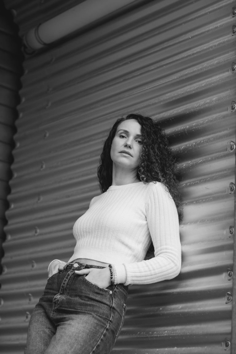 Portrait en noir et blanc d'une femme aux cheveux bouclés, vêtue d'un pull blanc et d'un jean, devant un mur métallique.