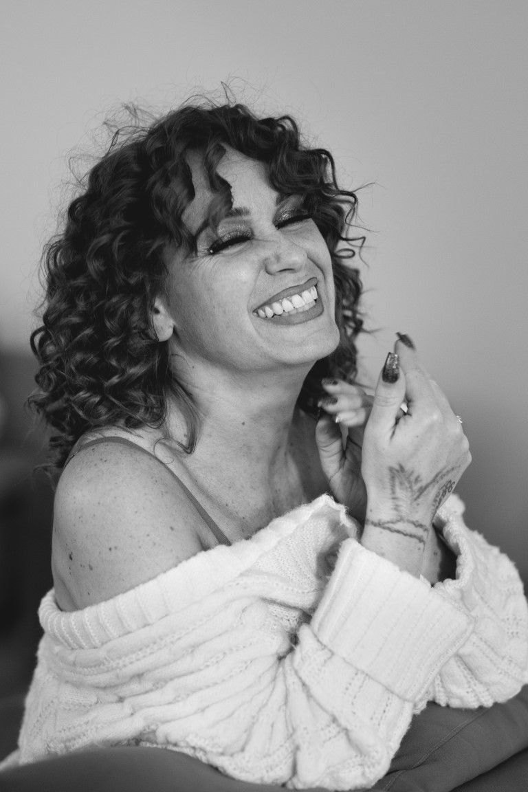 Black and white portrait of curly-haired woman laughing joyfully in off-shoulder knit sweater.
