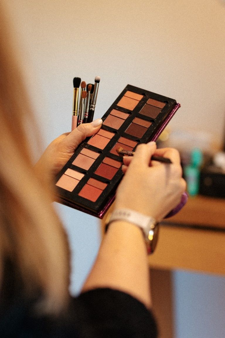 Woman holding an eyeshadow palette with warm brown tones and makeup brushes at a vanity table.