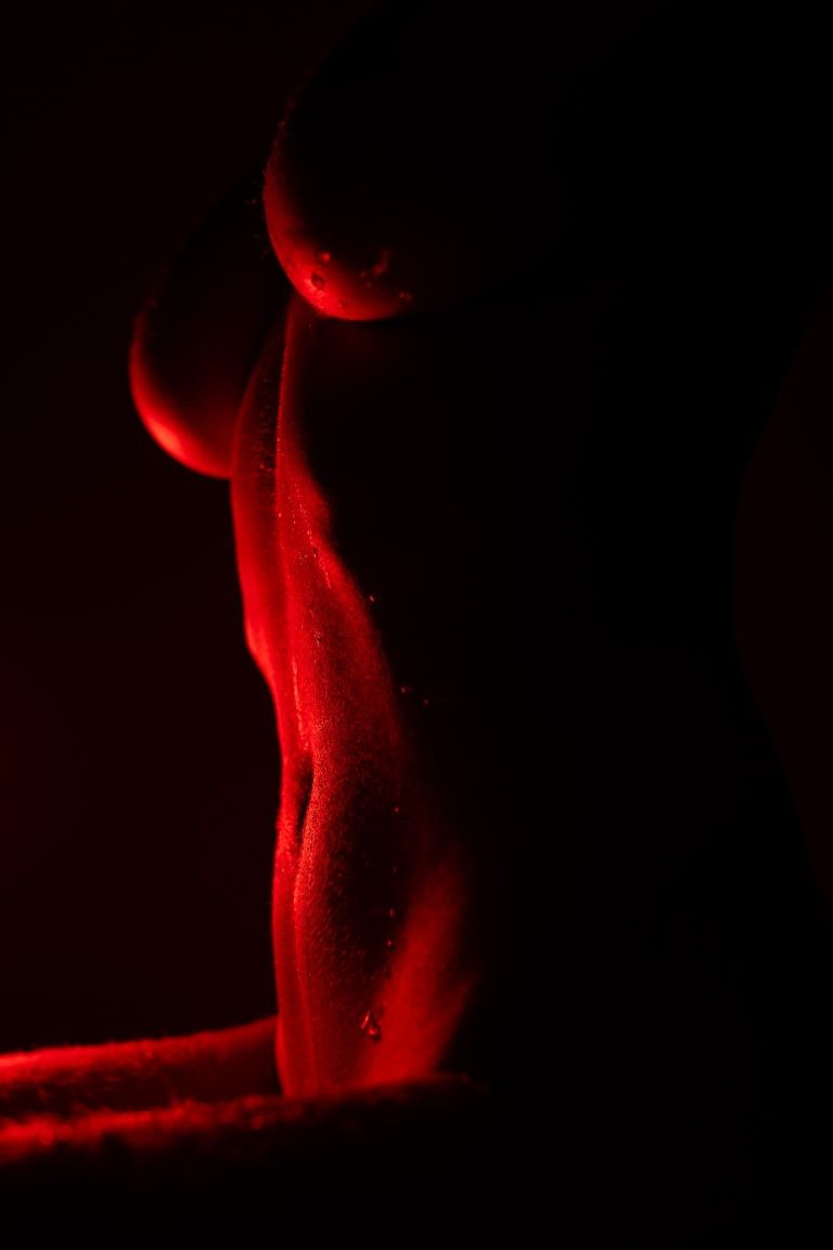 Silhouette artistique de nu éclairée en rouge, mettant en valeur le dos et les courbes d'une femme sur fond sombre.