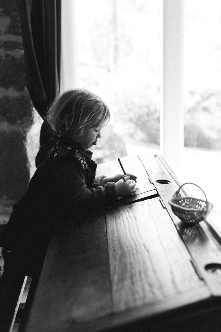 Jeune enfant assis à un bureau en bois dessinant près d'une fenêtre lumineuse, en noir et blanc.