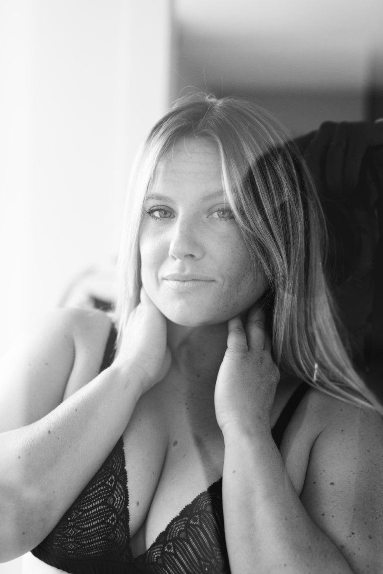 Black and white portrait of a blonde woman in lace top, hands on neck, gazing thoughtfully near a window.