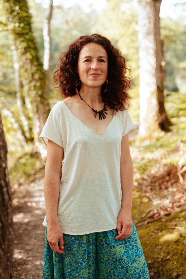 A woman with curly hair in a white top and blue floral skirt stands smiling in a sunlit forest.