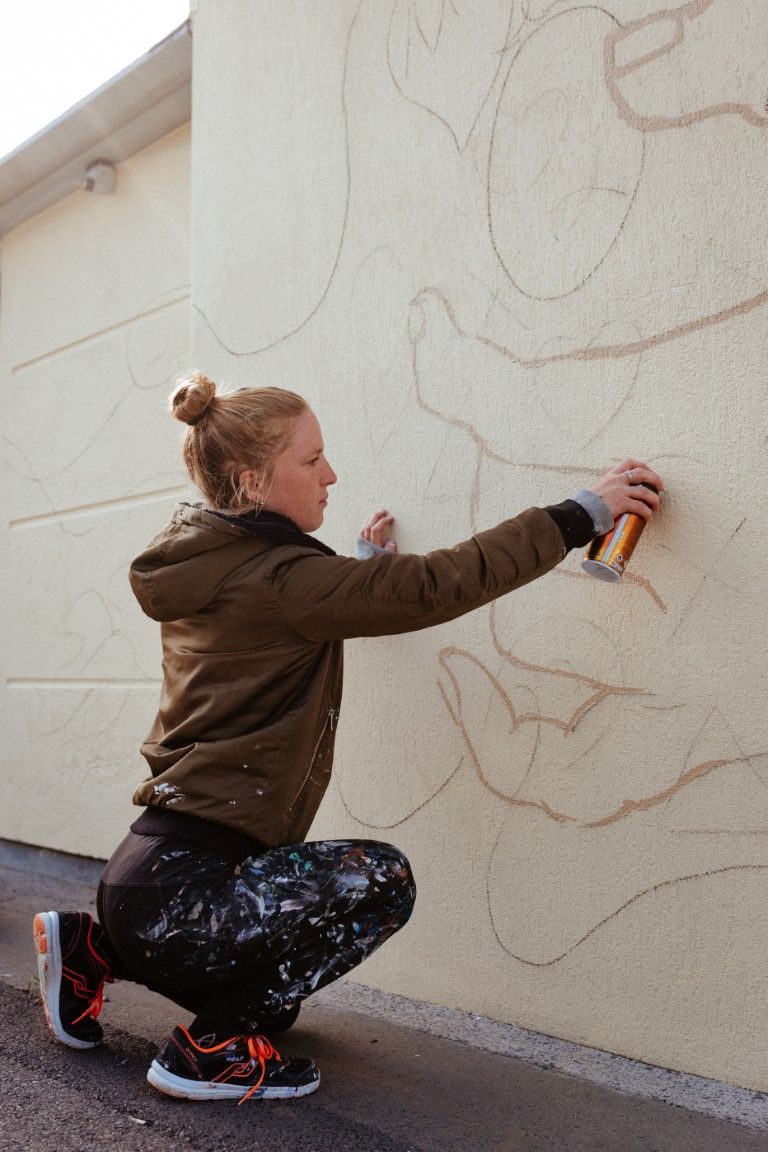 Woman crouching while spray painting outline sketch on exterior wall for street mural art.