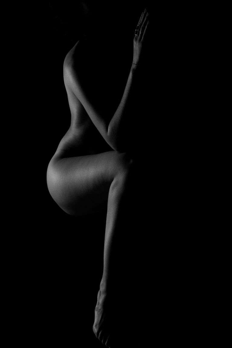 Black and white fine art nude silhouette of a seated figure against dark background, emphasizing curves and form.