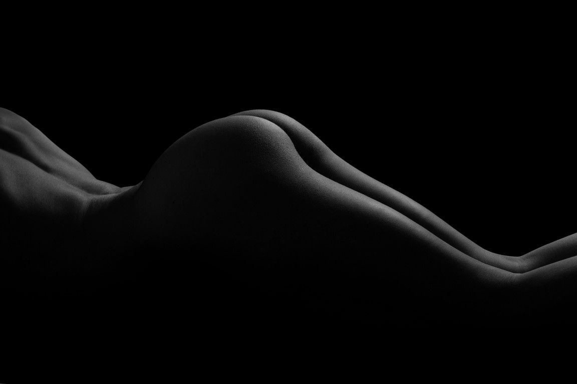 Black and white fine art nude photograph showcasing elegant body curves against a dark background.
