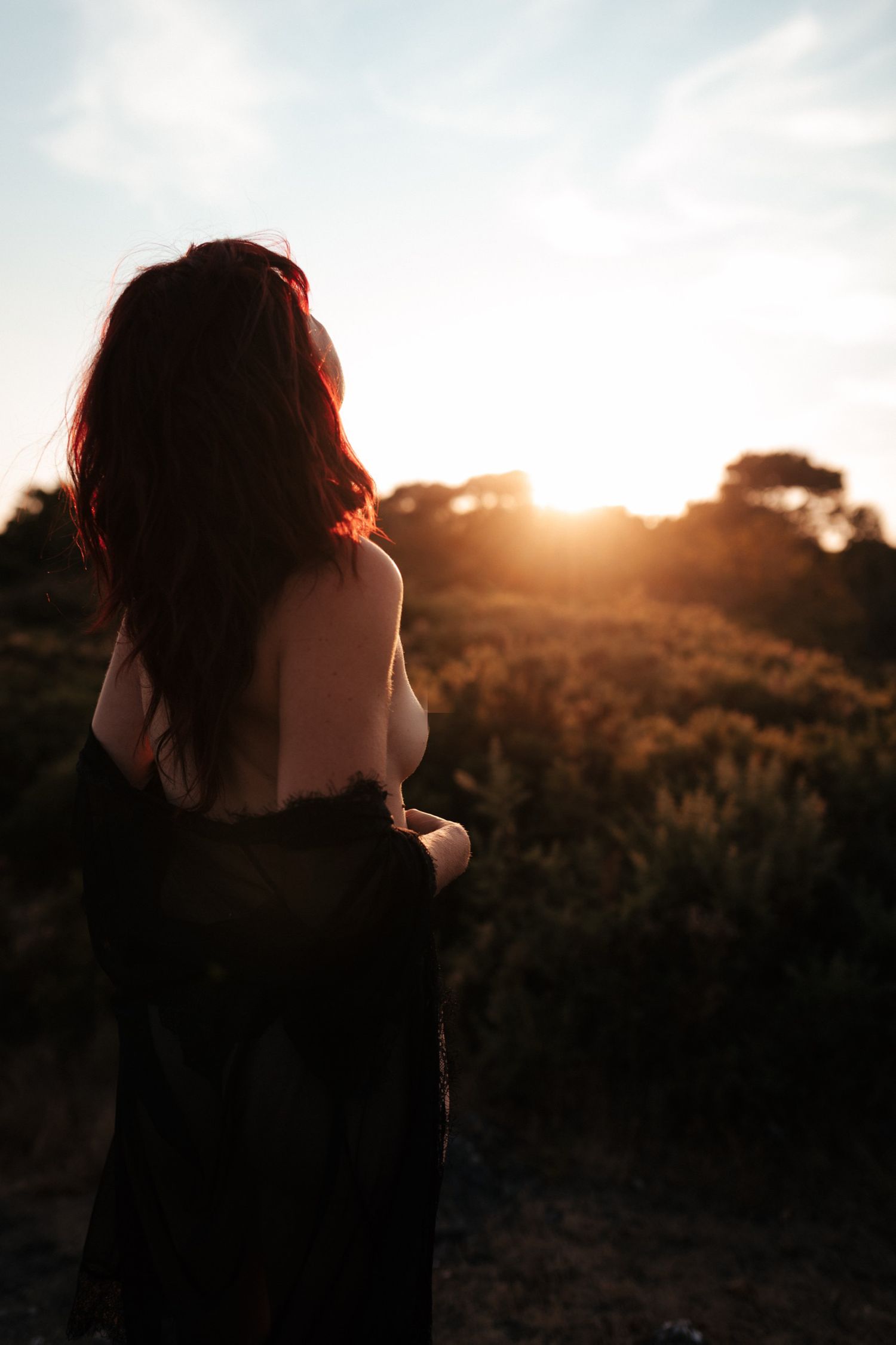 A woman with red hair stands facing a golden sunset over lush greenery, draped in a dark shawl.
