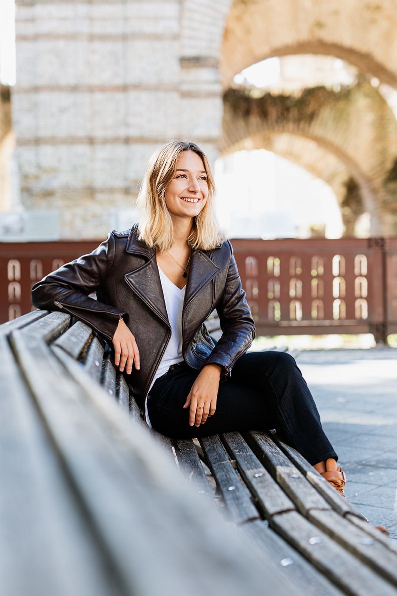 Young blonde woman in leather jacket sitting on bench outdoors near historic stone arch.