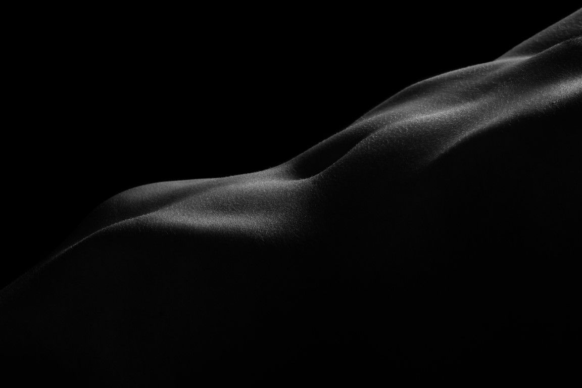 Black and white artistic bodyscape photograph highlighting curves and contours of the human form against dark background.