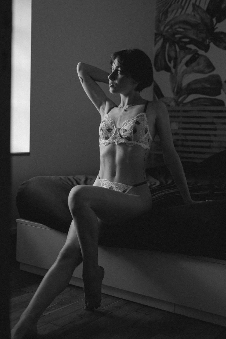 Black and white boudoir photo of a woman in white lace lingerie sitting on a bed.