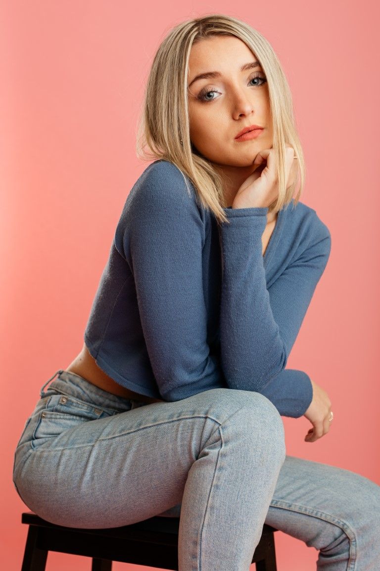 Femme blonde en pull bleu et jean assise sur un tabouret devant un fond rose.