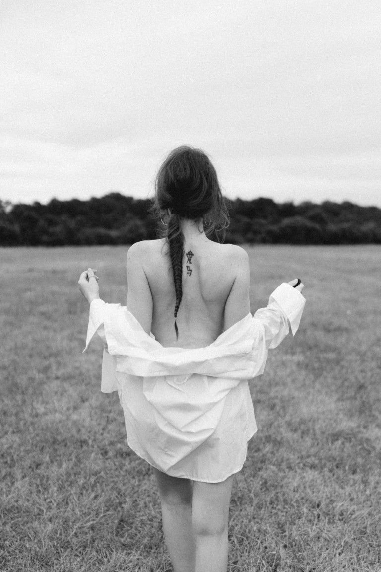 Vue arrière en noir et blanc d'une femme avec un tatouage sur la colonne vertébrale en train d'enlever une chemise blanche dans un champ.
