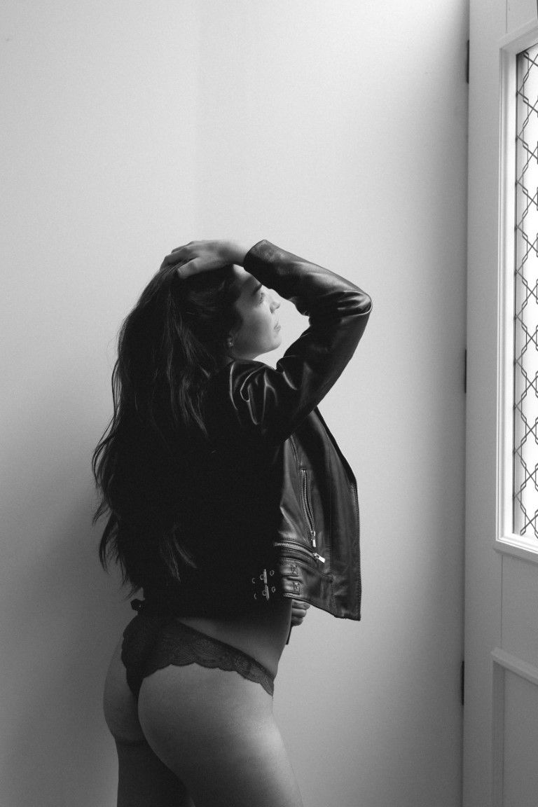 Black and white boudoir photo of a woman in lingerie and leather jacket posing by a window.