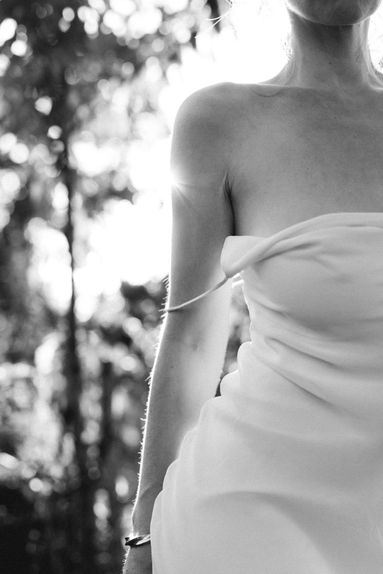 Black and white photo of a bride in a strapless white wedding gown standing outdoors among trees.