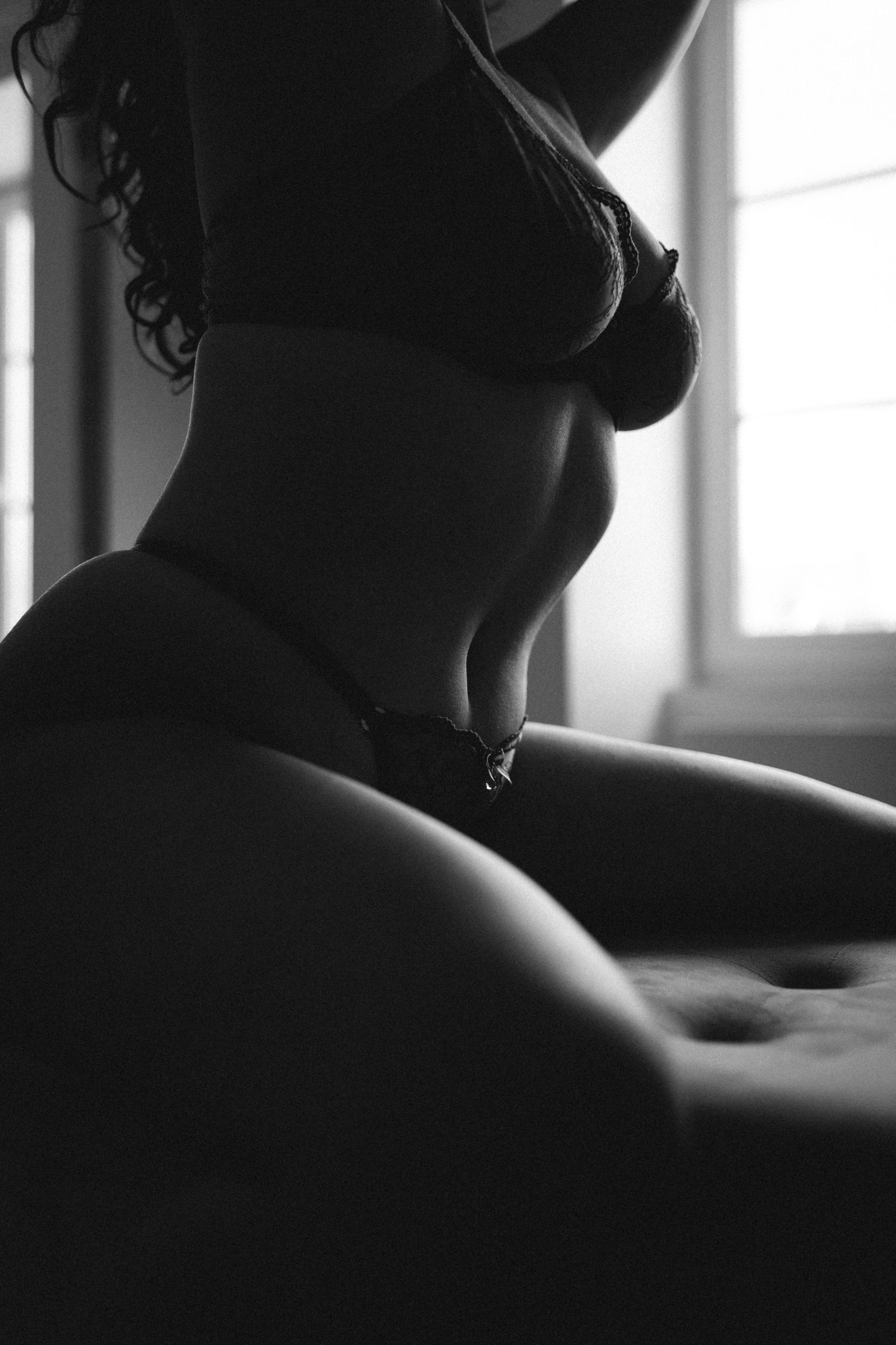 Black and white boudoir photo of a woman in lingerie posing by a window with dramatic lighting.