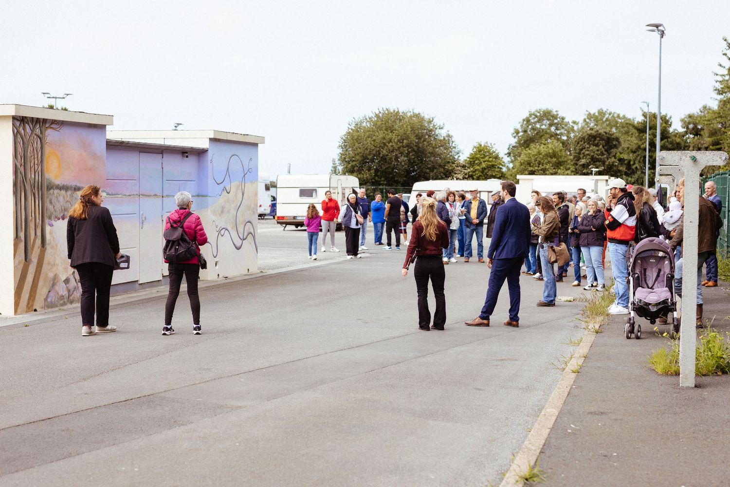 Groups of people gathered on a paved outdoor area near a mural wall on a cloudy day.