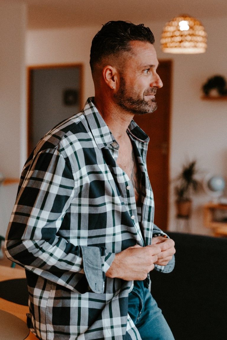 Un homme élégant, portant une chemise à carreaux en flanelle ouverte, regarde au loin dans un intérieur moderne.
