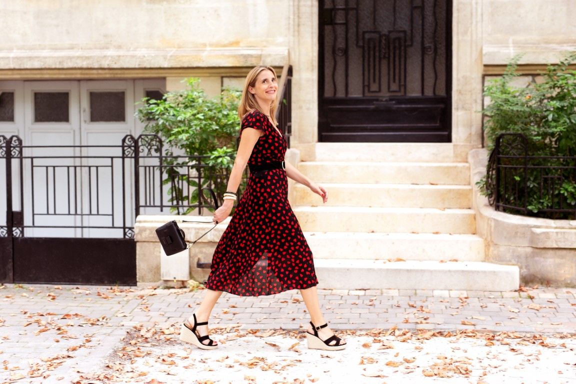 Blonde woman walking in red polka dot midi dress carrying handbag on cobblestone street.