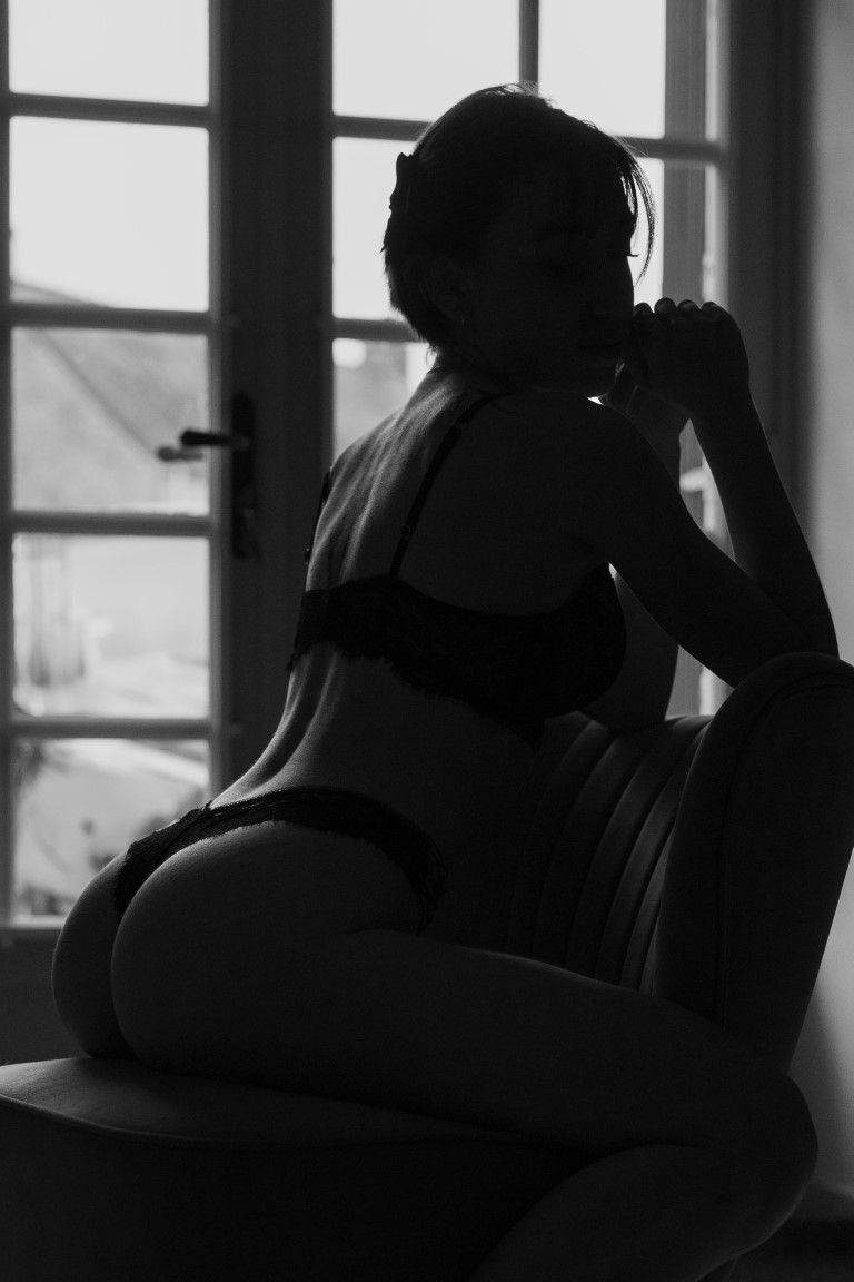 Silhouette of a woman in black lingerie sitting by a bright window.