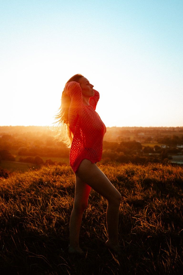 Woman in red mesh top poses on a sunlit hill at golden hour with hair blowing in the wind.
