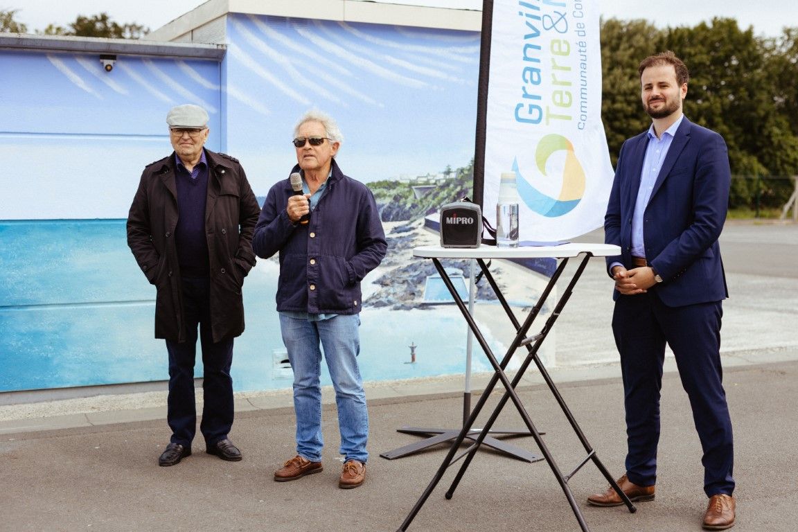 Three men stand outdoors at a Granville en Bord de Mer event, one holding a microphone near a display table.