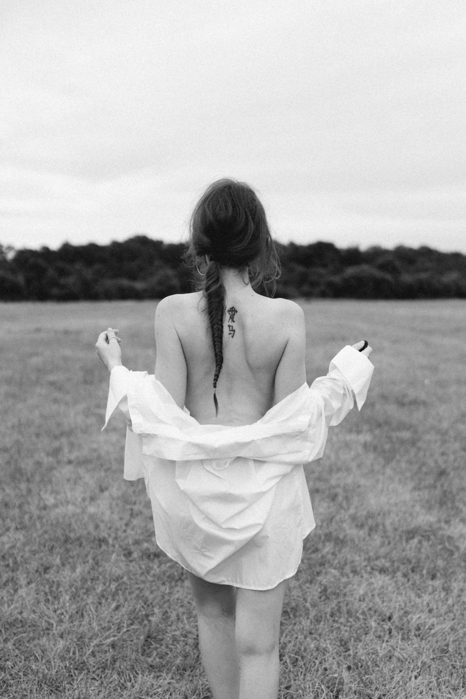 Black and white photo of a woman with a braid and spine tattoo, holding an open white shirt in a field.