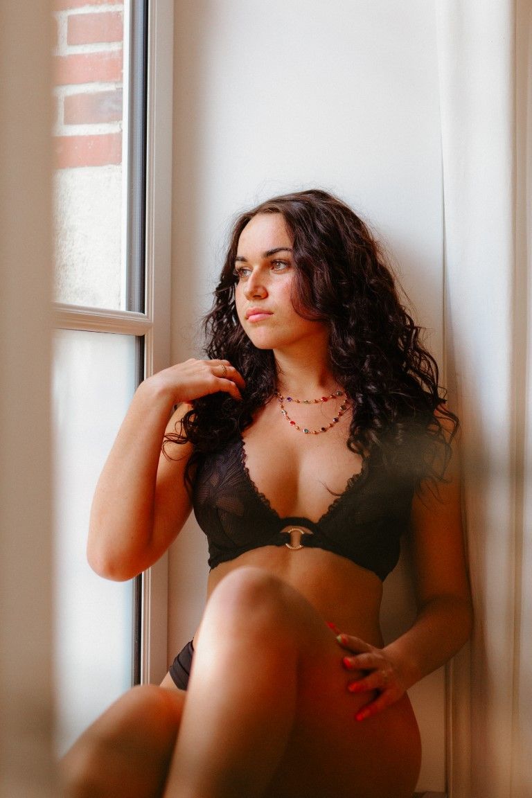 Woman in black lace bralette sitting by sunlit window.