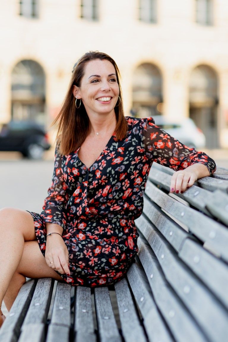 Woman in floral dress sitting on wooden bench outdoors, smiling confidently in urban setting.