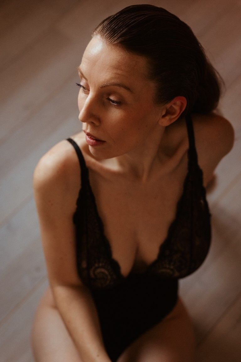 Femme en body de dentelle noire assise par terre, le regard détourné dans une lumière naturelle tamisée.