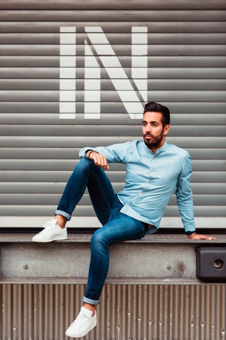 Un homme élégant et barbu, vêtu d'une chemise bleue et d'un jean, est assis décontracté contre un mur à volets métalliques.