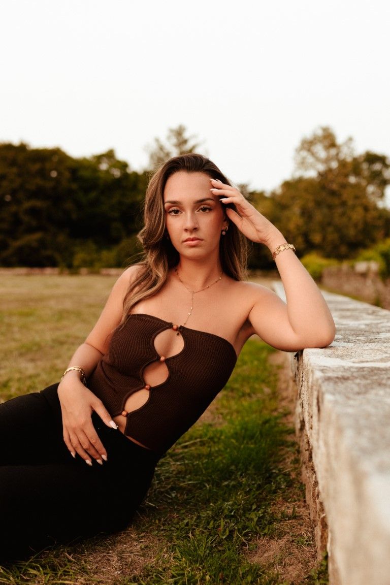 Jeune femme en robe noire bustier découpée posant devant un mur de pierre dans un parc extérieur au coucher du soleil.