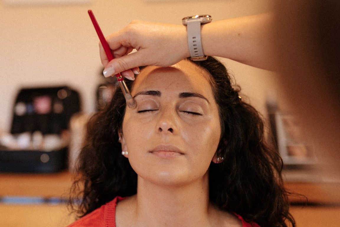 A makeup artist applying eyebrow makeup to a young woman with curly hair wearing a red top.