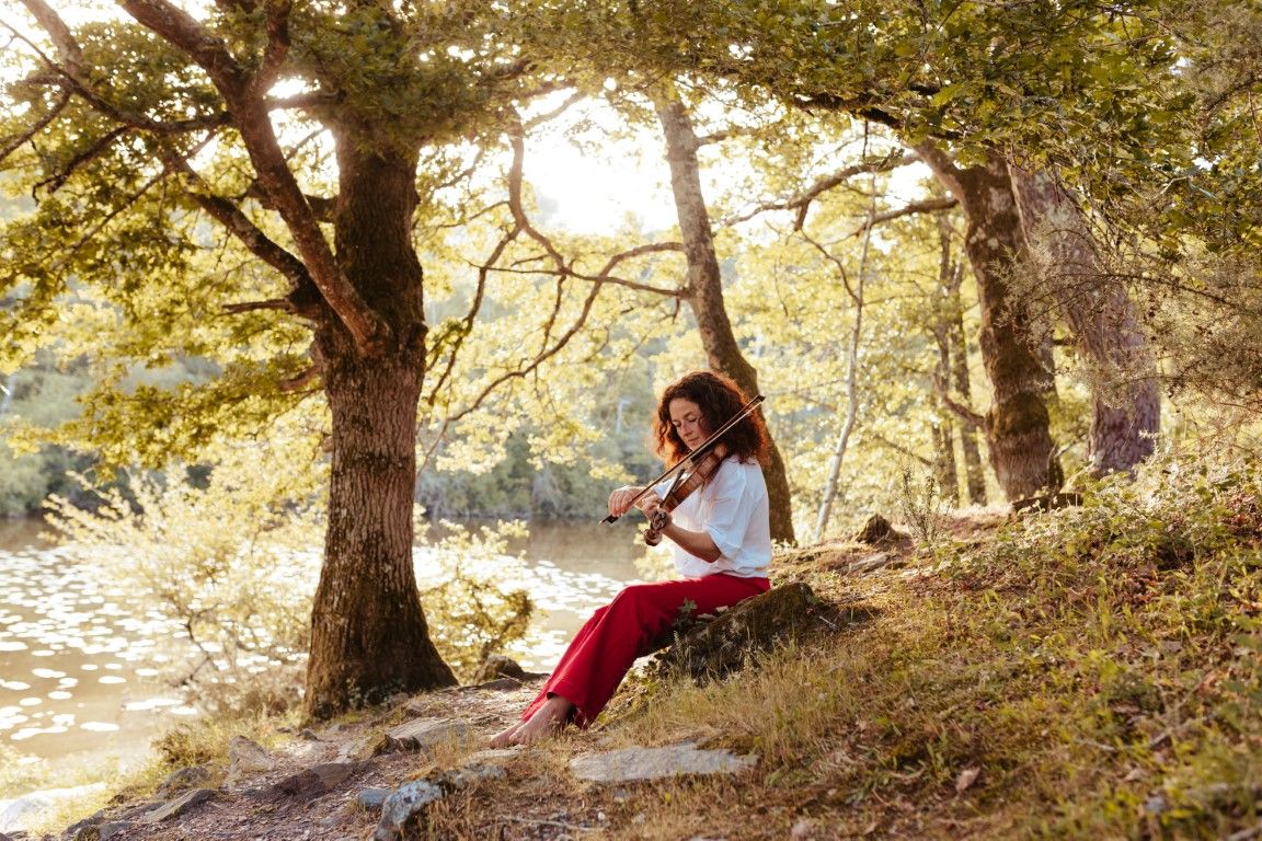 Woman playing violin outdoors by a sunlit river, sitting barefoot on a rock surrounded by lush green trees.