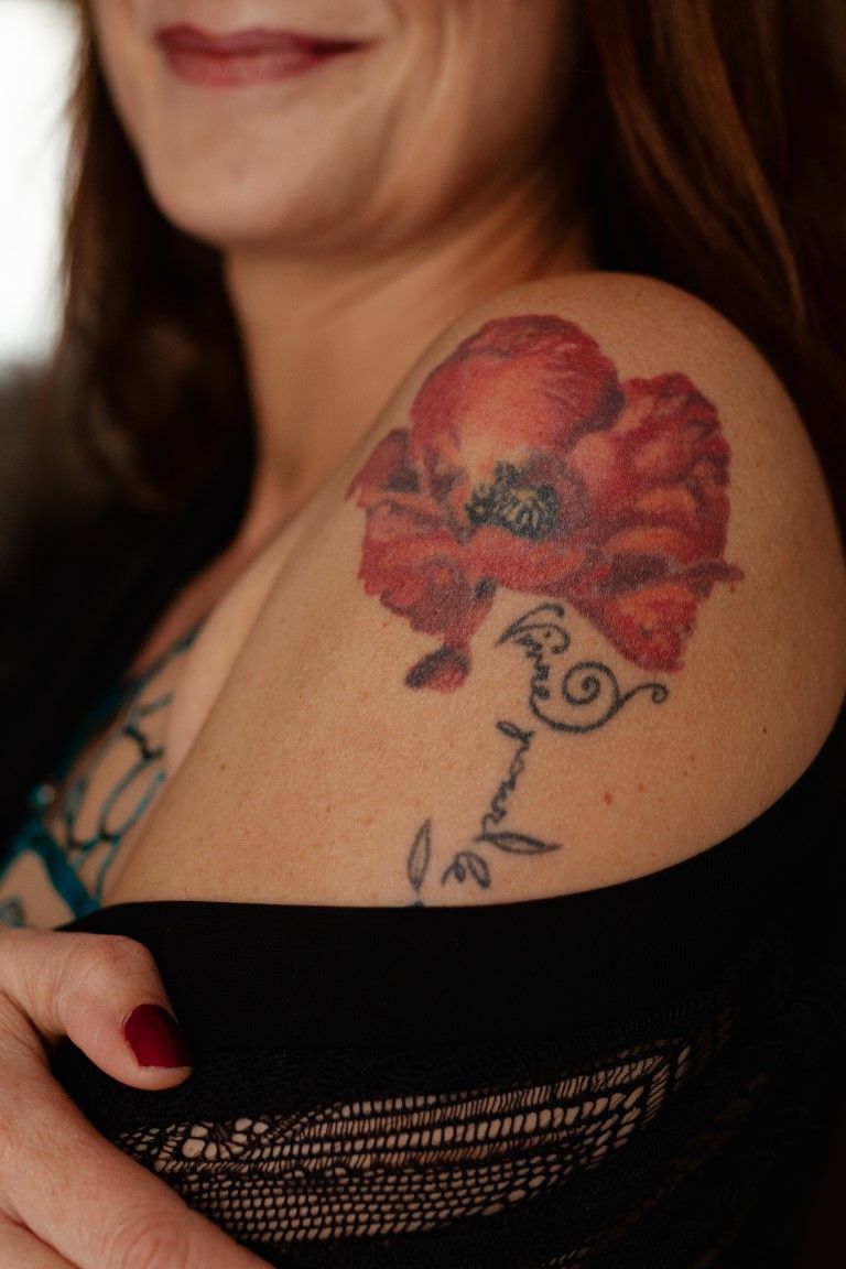 Gros plan sur un tatouage de coquelicot rouge vif sur l'épaule d'une femme, avec une inscription en écriture cursive.