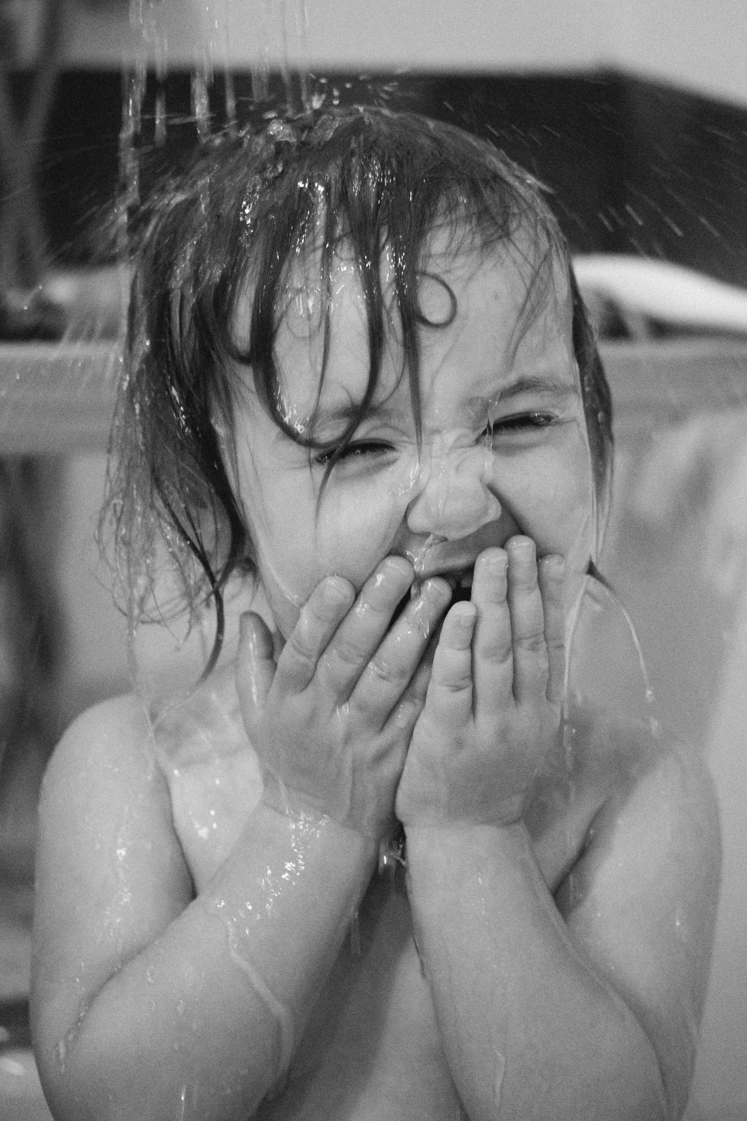 Photo en noir et blanc d'une petite fille qui rigole avec de l'eau qui lui coule sur le visage pour rincer son shampooing lors d'une séance