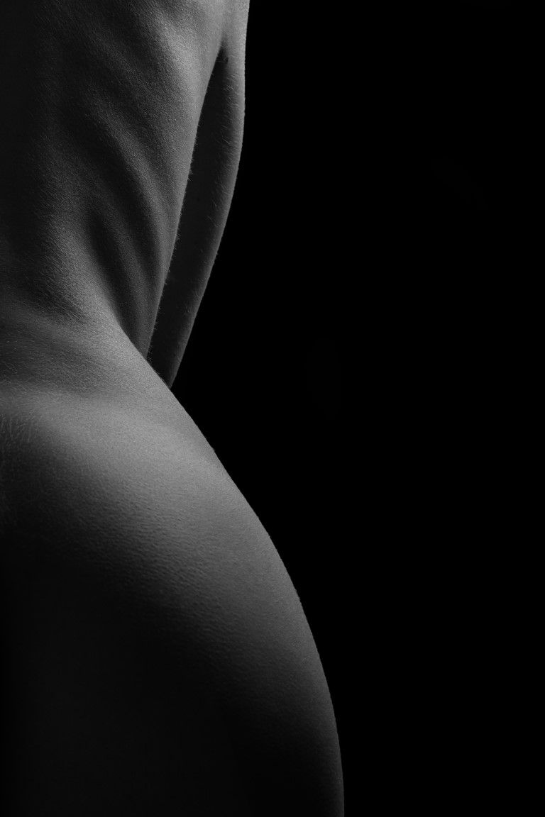 Black and white fine art nude photography highlighting the curves of the human back and hip against dark background.