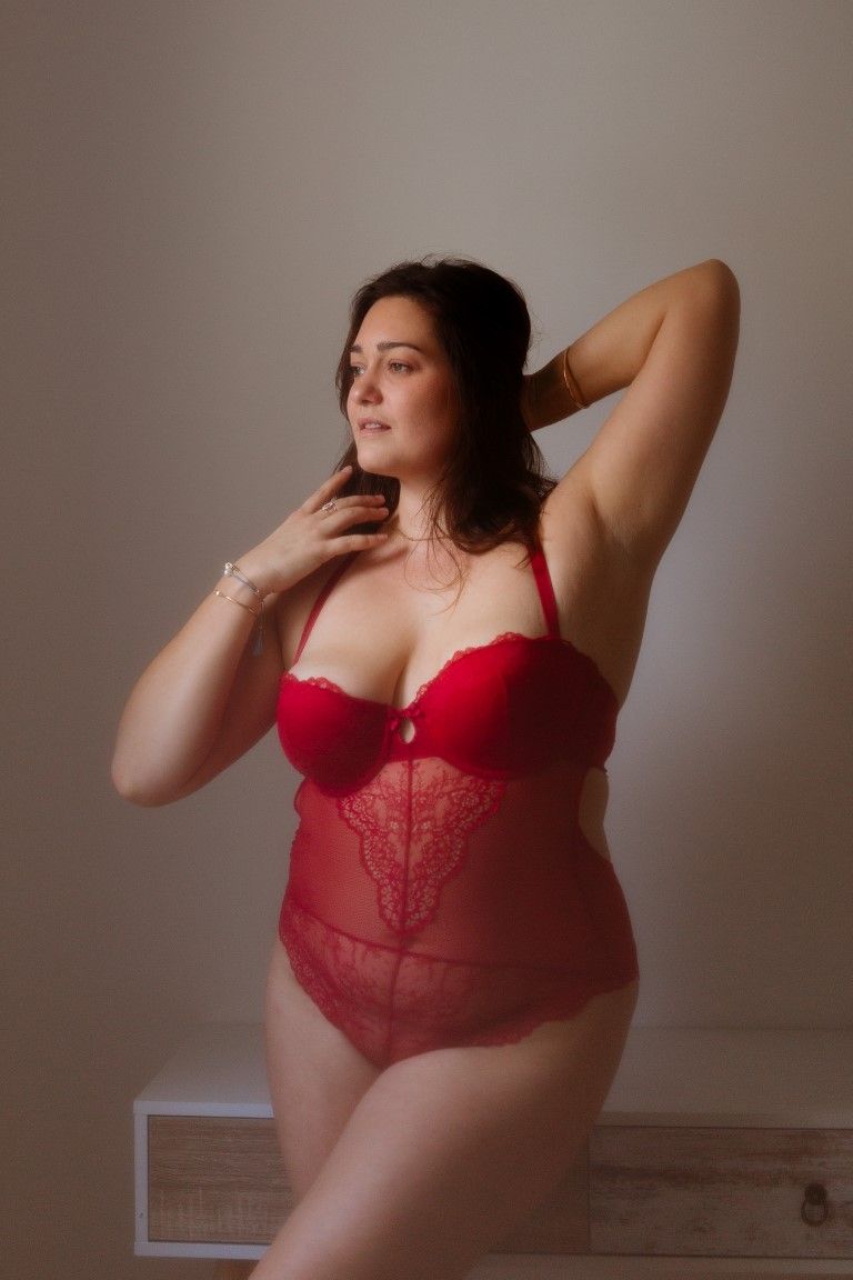 Curvy woman in red lace bodysuit posing with hands behind head against neutral background.