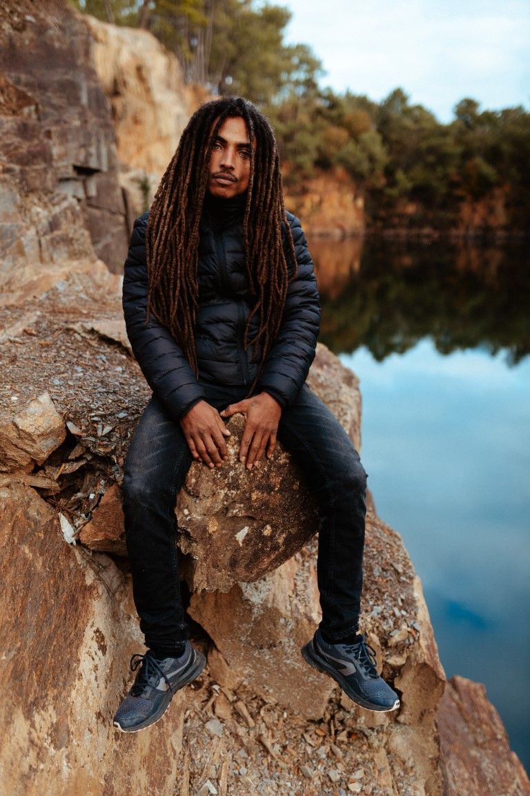 Un homme aux longues dreadlocks, vêtu de noir, est assis sur une falaise rocheuse au bord d'un lac calme et miroitant.